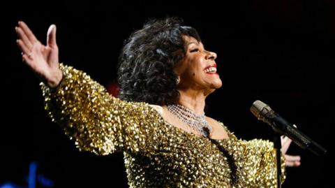 Dame Shirley Bassey performs on stage accompanied by The Royal Philharmonic Concert Orchestra during The Sound of 007 in concert at The Royal Albert Hall on October 04, 2022. She is wearing a gold sparkly dress and her arms are outstretched, she is smiling at the crowd. 