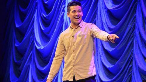 Tim Warwood stood on a purple-lit stage in a light pink shirt and black trousers. He is smiling and pointing and has short dark hair and is clean shaven.