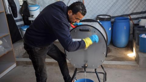 A man is turning a hand-cranked washing machine, which is a silver metal barrel with a tap at the bottom of it. Some water is coming out of the tap into a blue bucket. The man has dark hair and is wearing a navy fleece and black jeans. He is wearing blue and yellow rubber gloves. There are other barrels and buckets behind him, some with fabric in them.