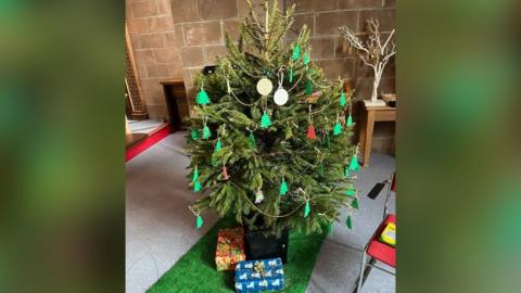 Medium-sized Christmas tree with white beads draped on it, Christmas tree-shaped baubles and some presents at the bottom.