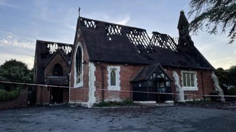 A brown and cream church that has been cordoned off and the roof has been burnt down. 