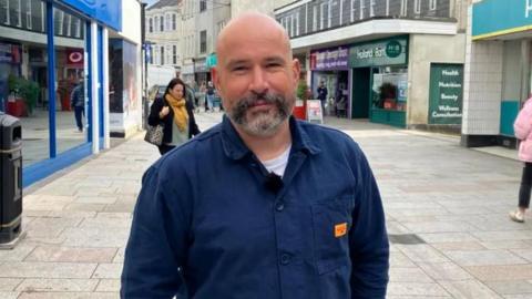 John Brown is a bald man with a dark grey beard, and is wearing a white T-shirt and navy blue overshirt. He is looking at the camera, smiling and has shops on either side of him while standing in Fore Street, St Austell.
