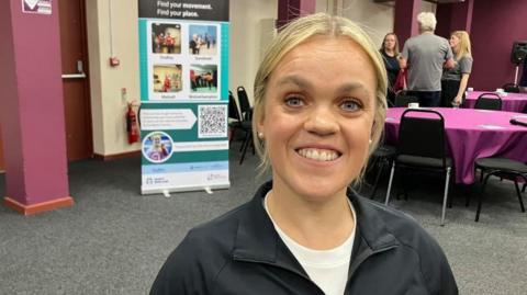A woman with blonde hair tied back. She is smiling and wearing a black sports top over a white T-shirt. She is standing in a room with grey carpet and circular tables. 