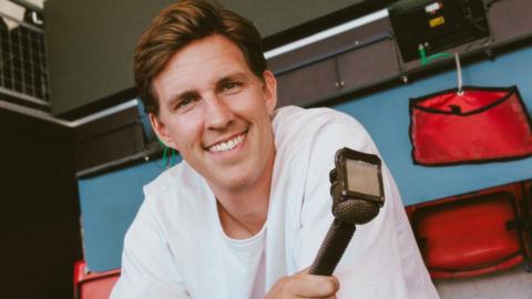 Joe Partington smiles for the camera. He is leaning on a desk in a football stadium. Behind him are red plastic seats. He is holding a radio microphone. He has short swept back brown hair and is wearing a white t-shirt.