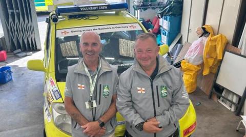 Two men stand in front of a bright yellow small car, with sirens fitted and sponsorship stickers. They are standing in a warehouse, with outboard motor, and a crash test dummy. They are wearing pale grey uniforms of Guernsey's St John volunteers, and smiling. 