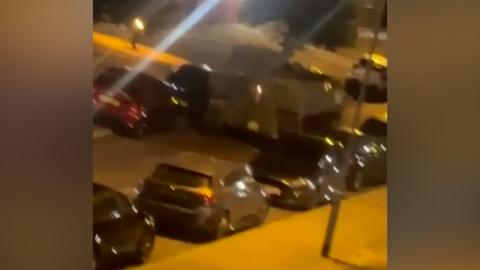 A blurry screencap from a video of a military vehicle being driven around Swinton Barracks in Wiltshire at night. The street is lined on both sides with parked cars and the military vehicle is being driven in between the cars. 