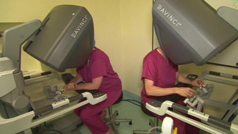 Two surgeons in maroon medical dress sit back-to-back with their heads in large grey consuls with the word 'Davinci TM' on them