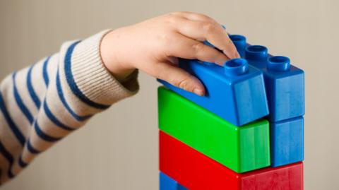 A child's hand placing a blue duplo brick on top of a multi-coloured tower of other duplo bricks.