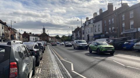 Yarm High Street has cars parked along both sides of the road on cobbles. 