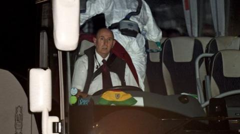 Andy Simonds sitting in the driving seat of the bus he drove, with a bucket Wales cap on the dashboard and people in PPE visible behind him