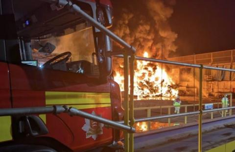 The fire burning at night with bright flames and a lot of smoke. A fire engine in the foreground and two fire fighters stand near the blaze.
