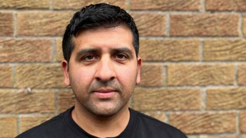 A picture of former cricketer Majid Haq. He is wearing a black T-shirt and is leaning against a brick wall, looking into the camera lens.