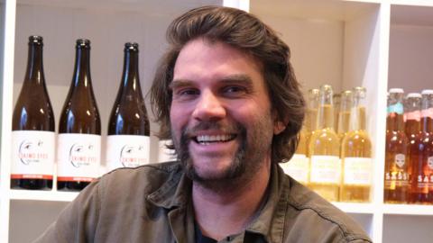 A man sits in front of a white shelf that is lined with bottles of cider. He has flowing dark hair with a large handle bar moustache.
