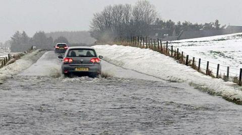 A car drives through floodwater covering a rural road, with snow piled along the roadside and fields under wintry cond