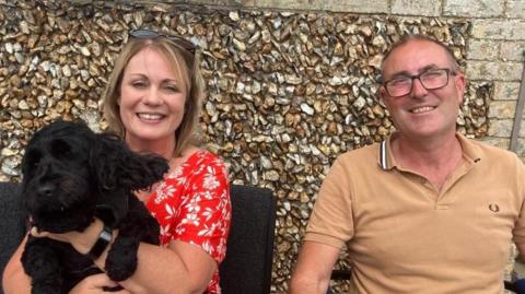 A woman on the left is holding a black dog, she is wearing a red top. On the right is a man wearing a light brown top, he is wearing glasses