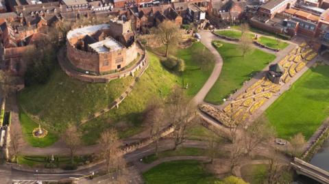 An aerial view of Tamworth Castle, a traditional motte and bailey style castle, sits on top of a mound with more modern buildings to one side and lawned areas to the other.