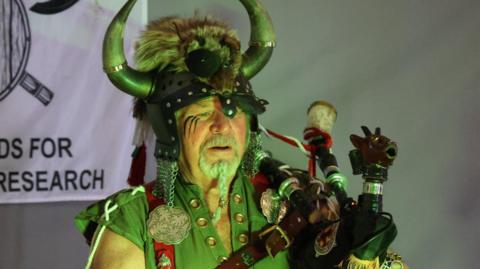 Terry Forster is standing on a stage, holding his bagpipes, and dressed as a Viking. He is wearing a helmet adorned with horns, fur and chainmail. He is wearing a green tunic with metal accessories on it in various Viking patterns. He is lit by a green light.