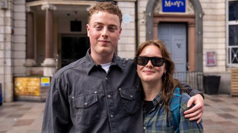 Joe, a man in a charcoal coloured denim shirt with a white tee underneath is stood next to Jess, a woman wearing black, tinted sunglasses and a chequered top. Joe has his hand around Jess; shoulders, and they stand on a street. 