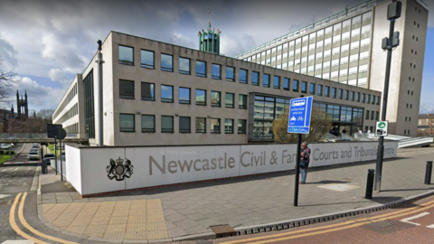 Newcastle Civil and Family Courts and Tribunals Centre is a white three-storey building with winows. Newcastle Civil and Family Courts and Tribunals Centre is written across a white fence alongside the city's Coat of Arms. A person is walkin on the pavement.