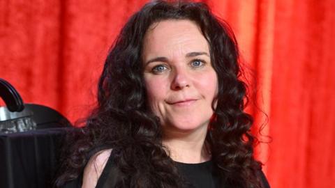 Cherylee Houston, with long black curly hair and wearing a black dress, sits in her wheelchair in front of a red curtain