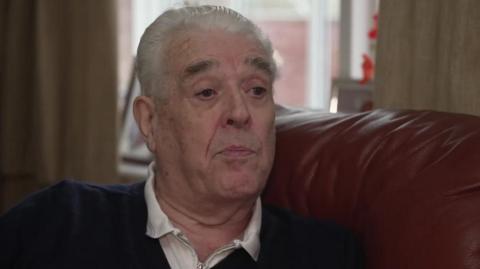 A man with short grey hair and a dark coloured jumper and white top is sitting on a burgundy sofa. 