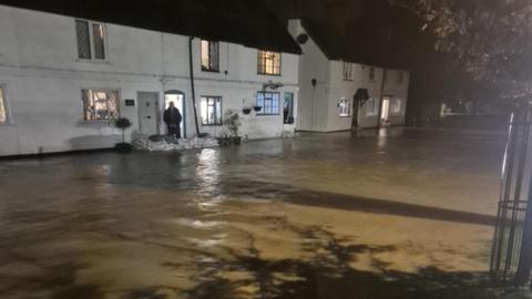 A number of white fronted old homes with sandbags in front with brown flood water flowing along the street.