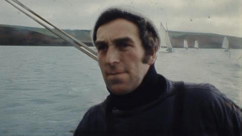 A grainy photo that shows Paul Laity on a boat looking past the camera. He is at sea and there are working boats under sail and a headland behind him. He has dark hair and is  wearing a woolly hat and a sou'wester. The sea is calm and the sky is grey with cloud.