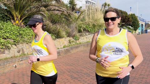 Two women in yellow tops with 'King's College Hospital Charity' on them run along the Eastbourne seafront.
