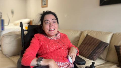 A woman in a wheelchair wears a red cardigan over a red and white chequered shirt. She wears a silver smart watch on her left hand and silver bracelet on he right. Behind her is a light brown sofa with dark brown pillows. 