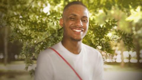 A smiling 20-year-old man in a white T-shirt standing in a park