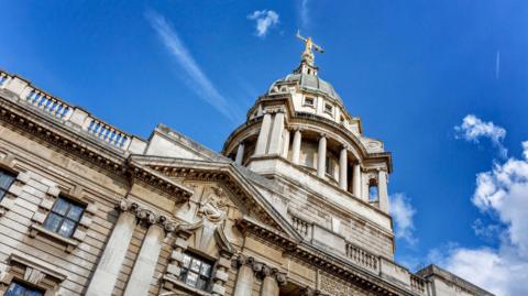 Exterior of the Old Bailey building looking upwards towards the Lady Justice on top of the dome with a blue sky in the background.