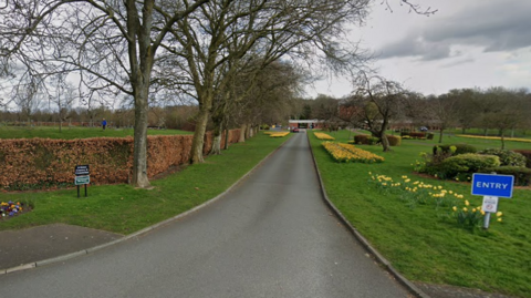 A Google Street View image of the road leading into St Helens crematorium. The road is lined on both sides by turf and flower beds.