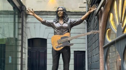 Bronze statue of Rory Gallagher with a guitar hanging off him, photographed against brick wall at Ulster Hall