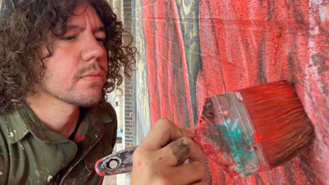 Head and shoulder shot of a street artist in khaki overalls, spattered with red paint holding a large decorator's paint brush and painting a brick wall