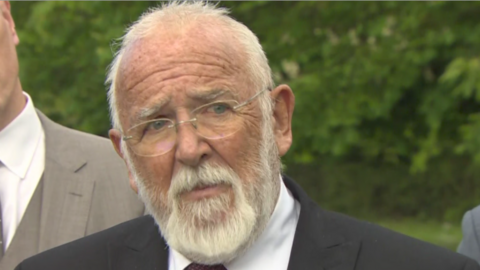 A man wearing a dark suit and white shirt stands in front of a camera. He has a short white beard and short white hair. He is wearing glasses.