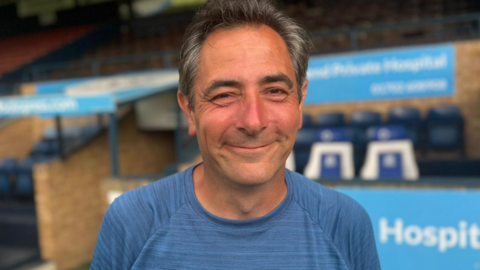 Karl Lansley, wearing a blue top and looking tanned after his marathon walk, smiles as he stands in front of one of the stands at Southend United's ground