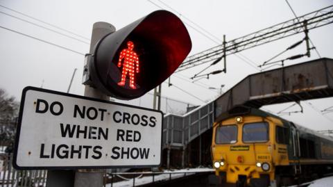 Signs at a level crossing, that shows a red person on a traffic light above a sign saying "do not cross when red lights show". A freight train crosses underneath a footbridge nearby.