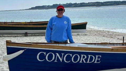 Klara Anstey standing on a beach next to a blue rowing boat that reads 'CONCORDE' on the side. She is wearing a red cap and a light blue jumper. The sea and two other rowing boats are behind her.