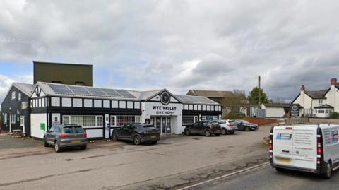 A black-and-white building which has a sign saying Wye Valley Brewery. Several cars are outside the building and a van is driving past. A white building with a grey roof and chimneys is in the background. 