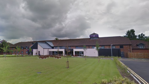 A two-storey hotel, with a large lawn and several picnic benches in front of the building. The purple Premier Inn branding is visible on what looks like some sort of tower on top of the roof