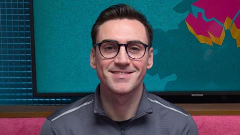 A man is looking straight at the camera. He has short jet black hair, which is slicked back. He has black-rimmed glasses. He is wearing a grey sports top with a zip. He is sitting in a podcast studio with a pink sofa, behind his head is a TV with the logo for The State of Us podcast, which is a teal background with a stylised image of Northern Ireland in pink.