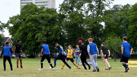Almost a dozen children are playing football on a field. Girls and boys are joining in and wear leggings, shorts and T-shirts, with some in blue vests to mark out the opposing team. There are large trees in the background.