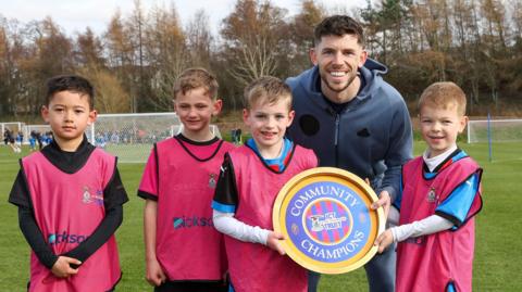 Ryan Christie na sheasamh le ceathrar bhalach agus iad air raoin-chluiche ball-coise. 