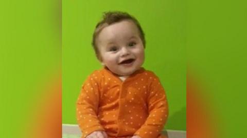 Undated image of the late baby Preston Davey. He has brown curly hair and brown eyes and is wearing an orange spotted baby grow. He is smiling. 