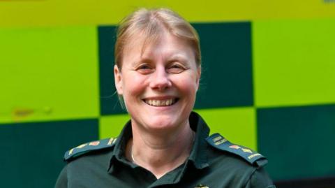 A smiling woman with blonde hair wearing a London Ambulance polo shirt with NHS and ambulance service logos on the front and epaulettes on the shoulders. An ambulance is in the background.  