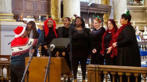 A conductor wearing a santa hat conducts a choir who wear Christmas jumpers and some have tinsel around their necks. All of the choir are women, wearing dark clothing and all are in a church with marble pillars and a wooden fence in front of them.