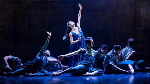 Eight ballet dancers dressed in blue swishy clothing under blue lighting. Most are low on the floor, one is lit more brightly and reaching upwards with one arm.