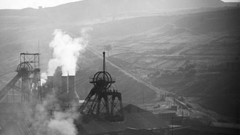 Black and white image of top side of a mining pit.  Smoke can be seen coming out of chimney pots.