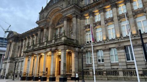 A browny/grey building with five columns at the front which are lit up. there are numerous windows on either side of the columns which go to three floors up. The building is ornate with stone arches above the columns. The sky is grey.