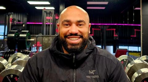 Ram Patten sitting in front of some dumbbells. He is wearing a black coat and is bald with a black beard